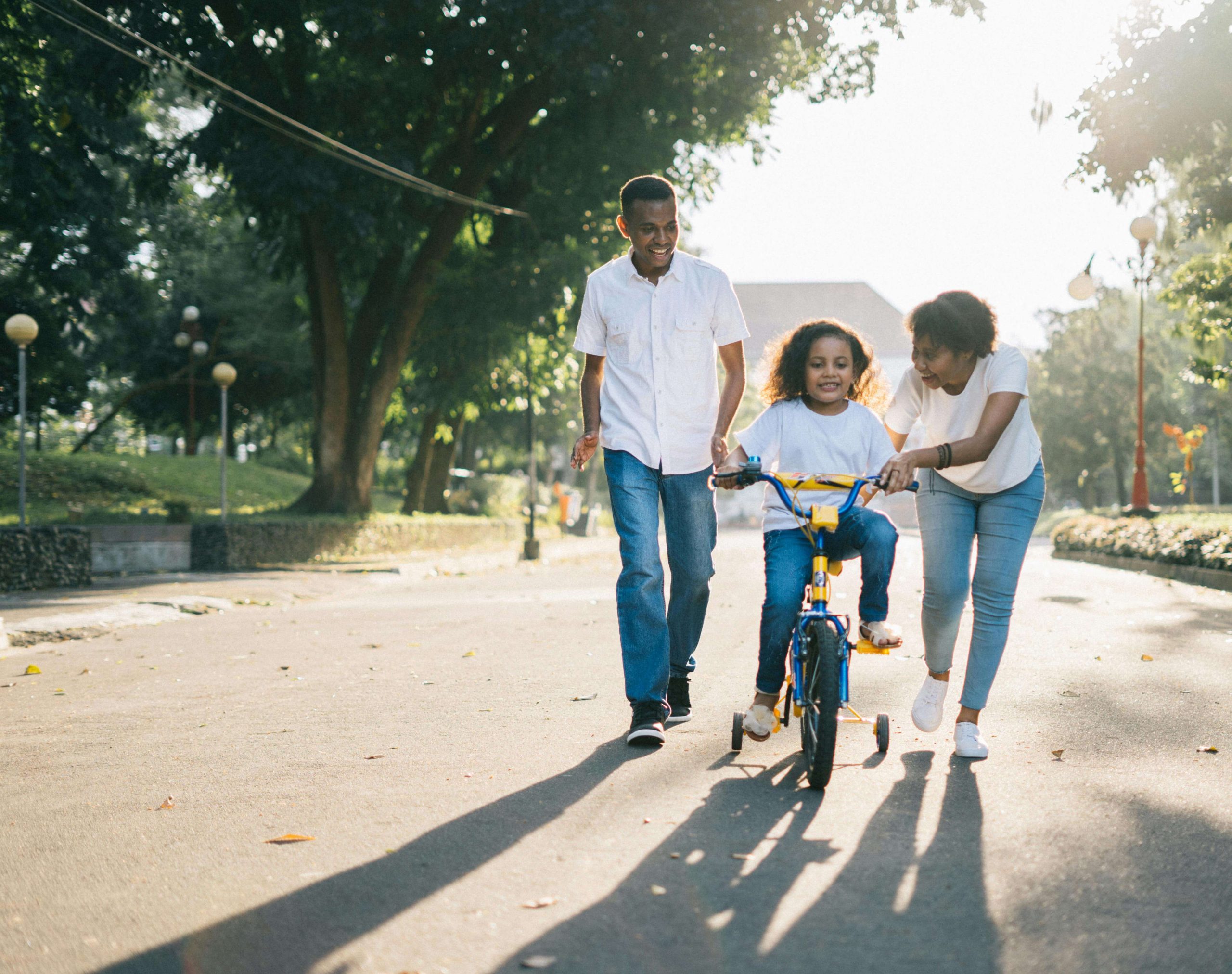 A child learning to ride a bicycle with assistance from two adults in a park.