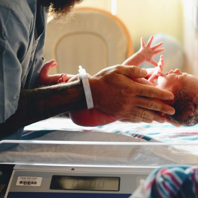 A newborn baby being carried to a weighing scale in a hospital.