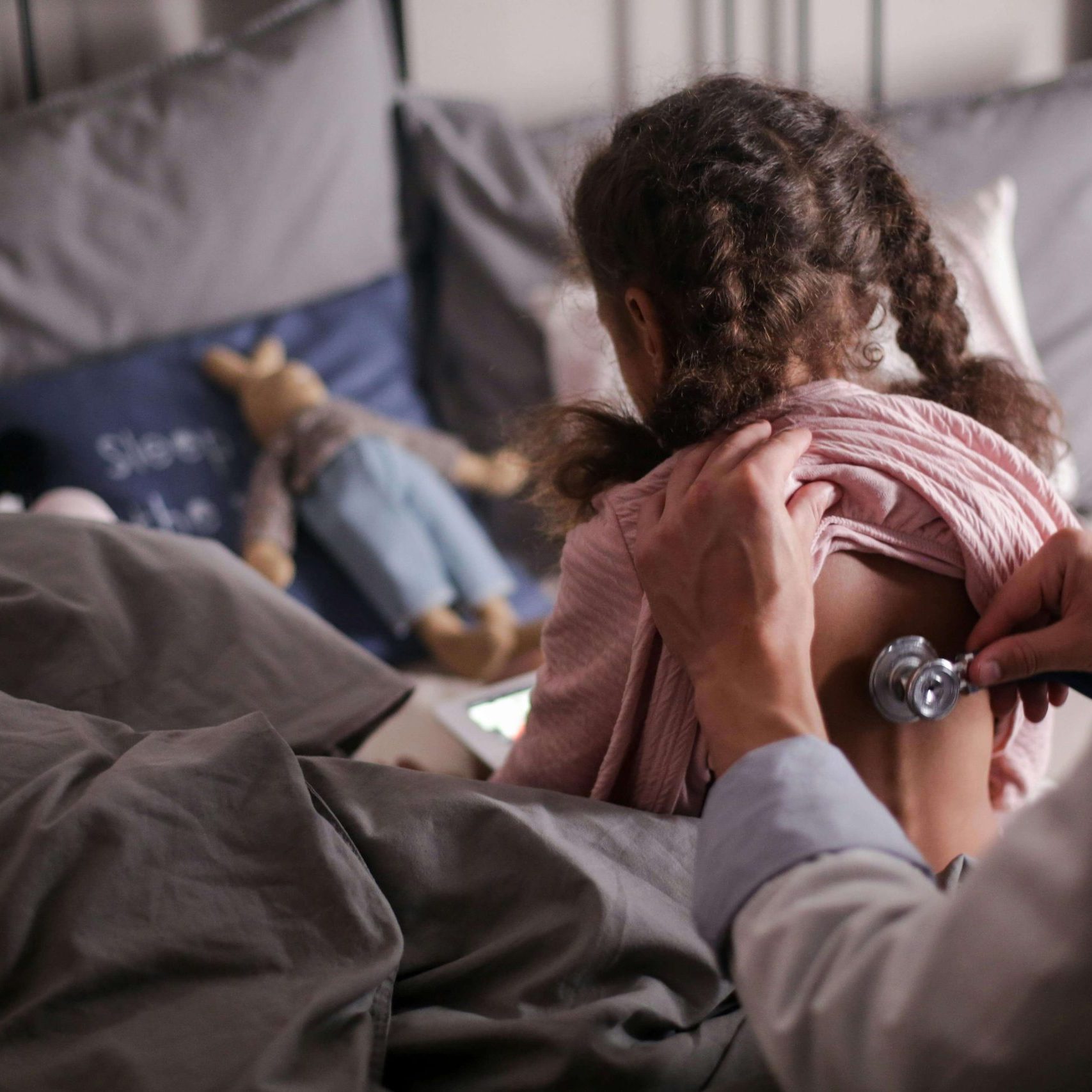 A child is sitting on a bed while a doctor uses a stethoscope on their back.