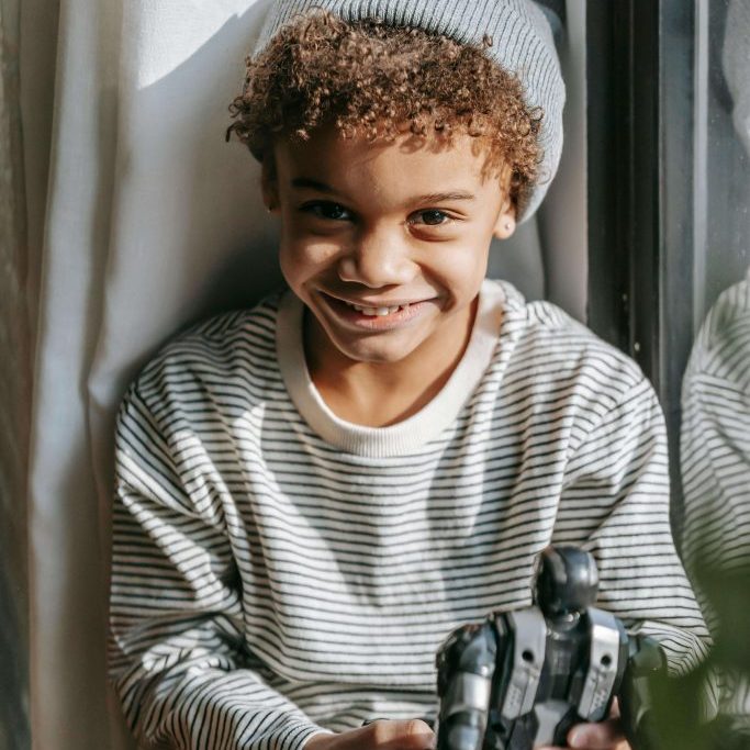 A child sits by a window, holding a toy.