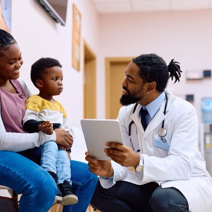 A doctor with a tablet conversing with a mother and her young child in a clinic.